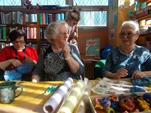 Panie siedzą przy stole i rozmawiają, trzymają gotowe pomponu, a bibliotekarka z tyłu wykonuje prace