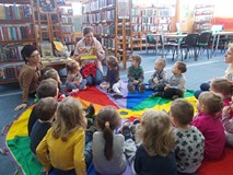 Dzieci siedzą na chuście wraz z bibliotekarką, która pokazuje im książeczkę