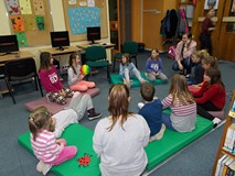 Dzieci siedzą na materacach przed bibliotekarką, a dziewczynka trzyma w ręku piłkę