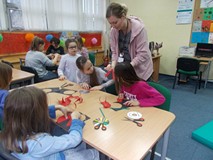 Dzieci siedzą przy stole i wykonują prace plastyczną, bibliotekarka pokazuje im jak ją wykonać