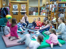 Dzieci z bibliotekarką siedzą na materacach, a dziewczynka rzuca piłką