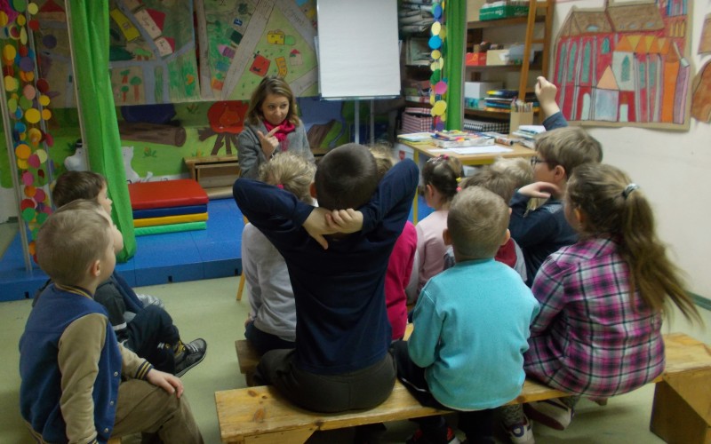 Grupa przedszkolaków siedzi na ławeczkach, a pani bibliotekarka czyta wiersze.