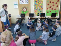 Dzieci z rodzicami i bibliotekarką z opaskami i ilustracjami na głowach siedzą na dywanie i bawią si