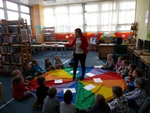 Dzieci siedzą na chuście obserwując bibliotekarkę, która recytuje wiersz z piórkiem w ręku