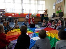 Dzieci siedząc na chuście słuchają opowiadania czytanego przez bibliotekarkę, która trzyma w ręku za