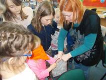 Bibliotekarka trzyma pudełko, z którego dzieci z zasłoniętymi oczami losują rekwizyty
