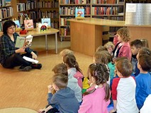 Dzieci siedzą na chuście integracyjnej i słuchają bibliotekarki czytającej książkę o 