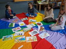 Dzieci siedzą na chuście na której rozłożone są ilustracje i zgłaszają się do odpowiedzi