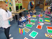 Dzieci siedzące wokół gry planszowej patrzą na bibliotekarkę, która wyjaśnia im zadanie