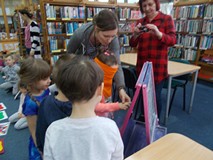 Bibliotekarka pomaga dorysowywać trąbę słonia dziewczynce, która ma zasłonięte oczy