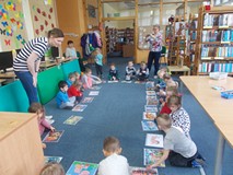 Dzieci siedzą wokół kolorowych ilustracji, a bibliotekarka wyjaśnia im zasady zabawy