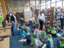 Dzieci siedzą na ziemi z obrazkami w ręku, a bibliotekarka rozdaje im cukierki