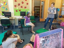 Dziewczynka siedzi na krześle z ilustracją na głowie, a reszta dzieci i bibliotekarka odpowiadają na