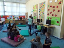 Dzieci siedzą na materacach podczas gdy bibliotekarka wyjaśnia dzieciom zasady zabawy