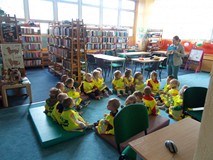 Dzieci siedzą na materacach przed bibliotekarką, która pokazuje im książkę