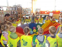 Dzieci trzymają chustę animacyjną z kolorowymi balonami w środku, a bibliotekarka objaśnia im zasady