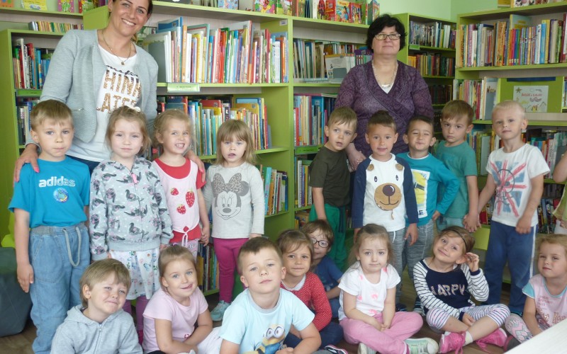 Dzieci stoją przy regałach ze swoją wychowawczynią i bibliotekarką