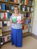 Pracownica Działu Opracowania Teresa Miękus stoi na tle regalu z książkami z polecanymi przez siebie