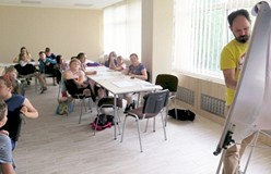 Dzieci siedzą przy stole, a pan prowadzący warsztaty komiksowe rysuje na tablicy