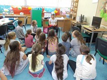 Bibliotekarka wraz z dziećmi siedzą naokoło dziewczynki, która siedzi na krześle z ilustracją zwierz