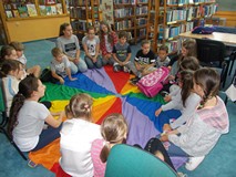 Dzieci siedzą na chuście animacyjnej, a dziewczynka wyjmuje rzeczy z różowego plecaka