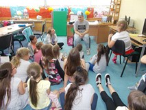 Dziewczynka siedzi na krześle z ilustracją na głowie, a reszta dzieci z bibliotekarką pomagają jej o