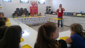 Dzieci siedzą przy stołach słuchając prowadzącego warsztaty chemiczne