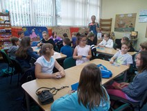 Dzieci siedząc przy stołach słuchają bibliotekarki