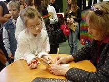 Przy stoliku siedzi pani Wanda Chotomska i podpisuje książkę pochylonej na stolikiem dziewczynce po 