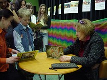 Przy stoliku siedzi pani Wanda Chotomska i podpisuje książkę stojącemu przy stoliku chłopcu po spotk