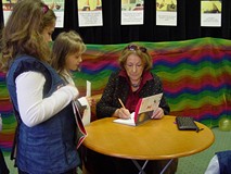 Przy stoliku siedzi pani Wanda Chotomska i podpisuje książkę stojącym po lewej stronie stołu dwóm dz