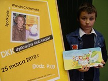 Chłopiec pozuje z podpisaną książką pani Wandy Chtomskiej obok zółtego plakatu promującego spotkanie
